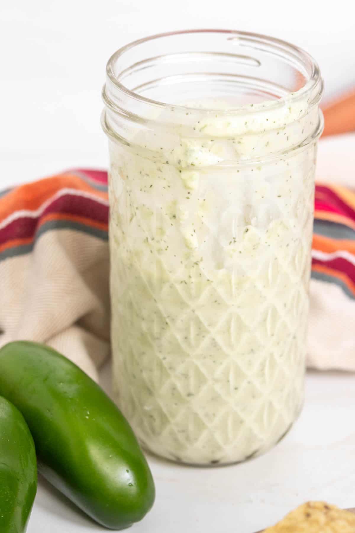 A jar filled with a creamy, white sauce with visible herbs. The jar is on a surface next to two green jalapenos and a striped cloth.