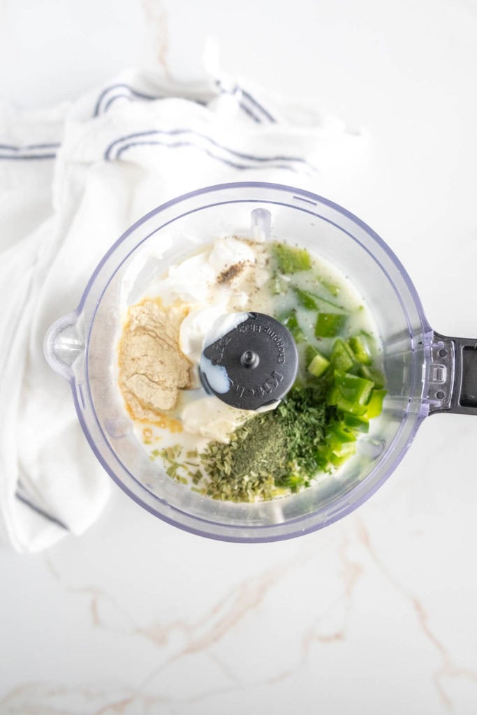 A food processor bowl containing various ingredients, including green peppers, herbs, a creamy substance, and powdered spices, set on a marble countertop. A white cloth with blue stripes is nearby.