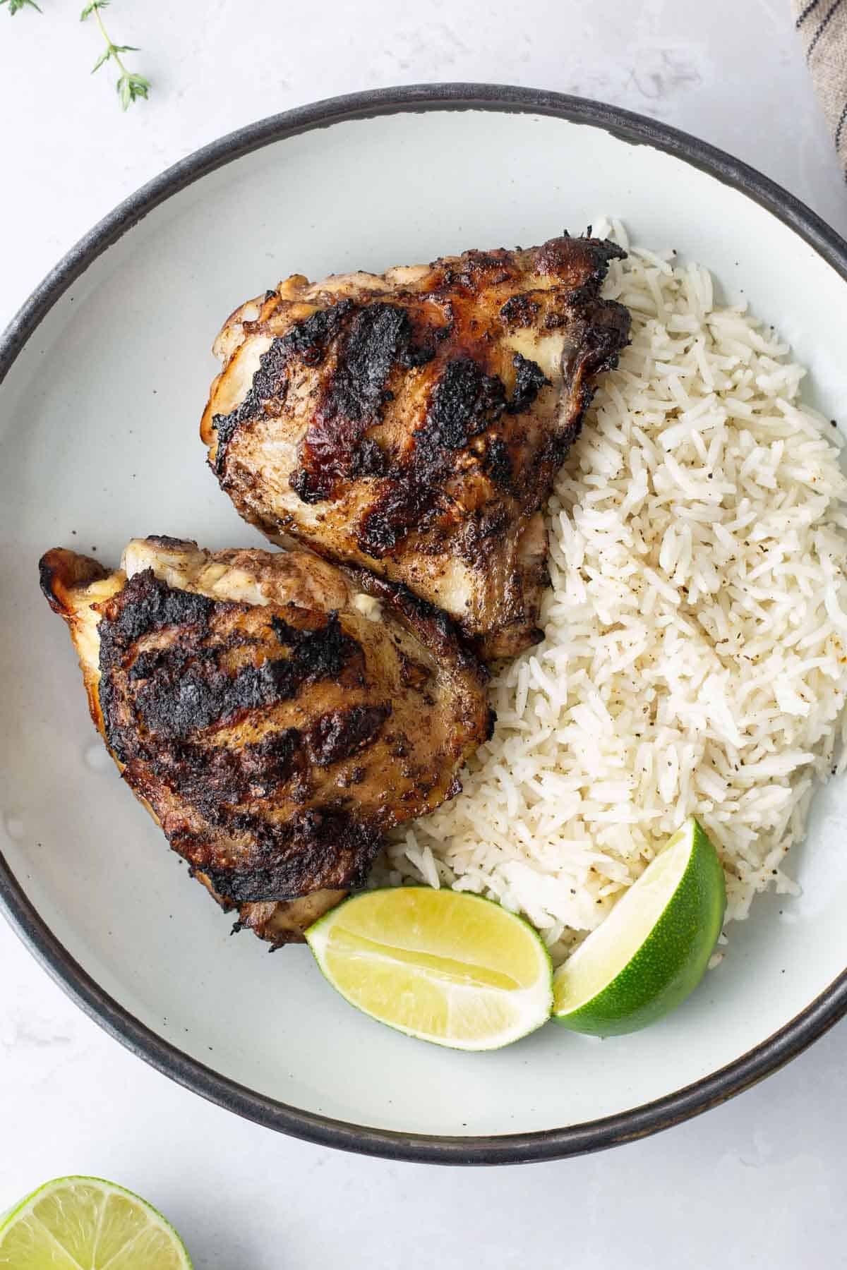 A plate with two grilled chicken thighs, a serving of white rice, and two lime wedges, set on a white surface.