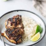 A plate with grilled chicken, white rice, and lime wedge. Fork on the side, whole limes, and striped cloth visible in the background.