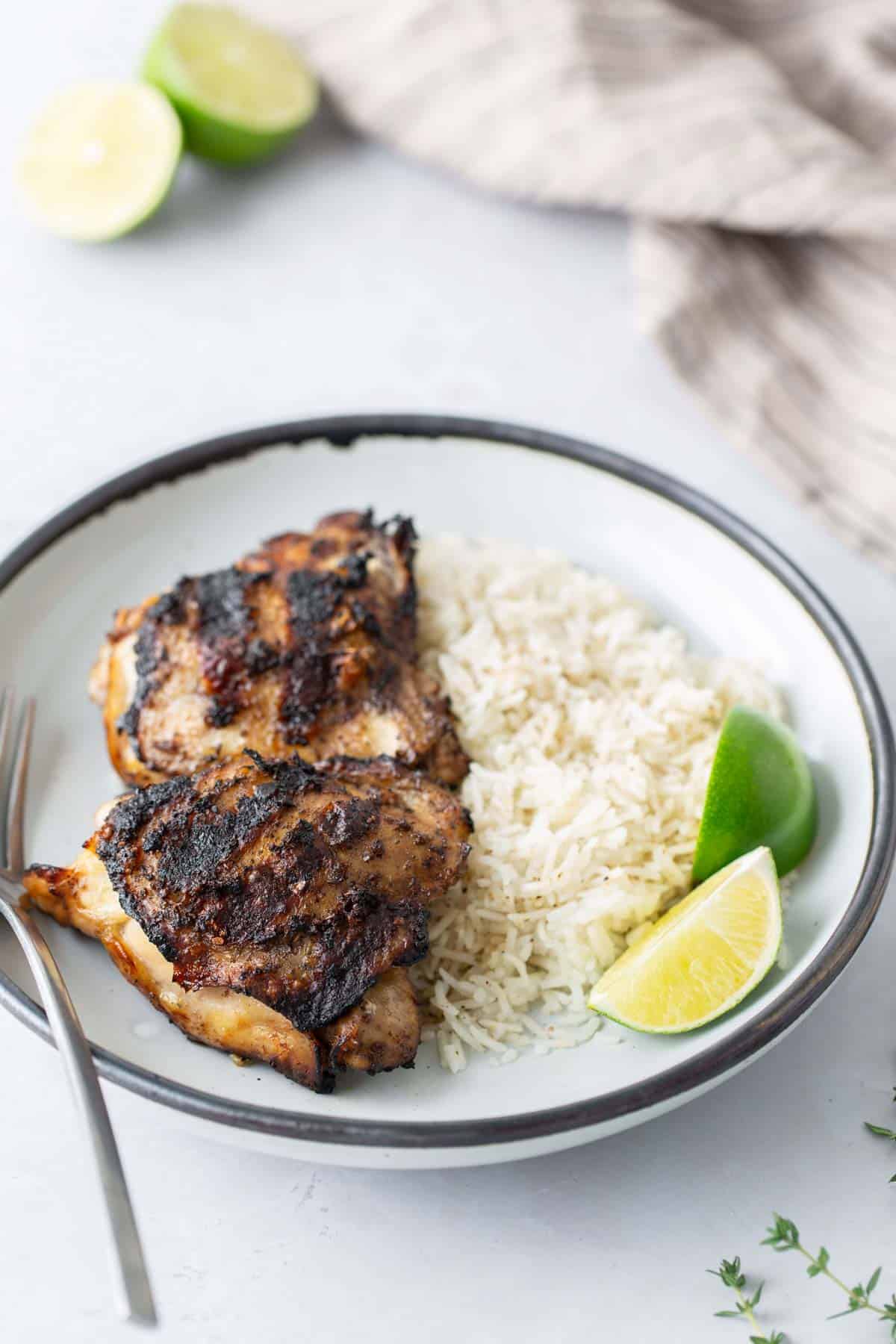 A plate with grilled chicken, white rice, and lime wedge. Fork on the side, whole limes, and striped cloth visible in the background.