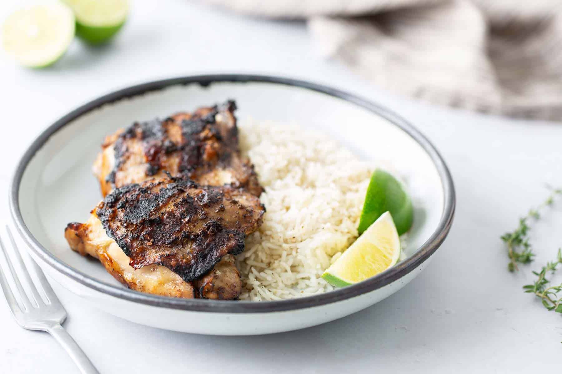 A plate containing grilled chicken, white rice, and lime wedges. A fork is placed beside the plate, and a gray napkin is in the background.