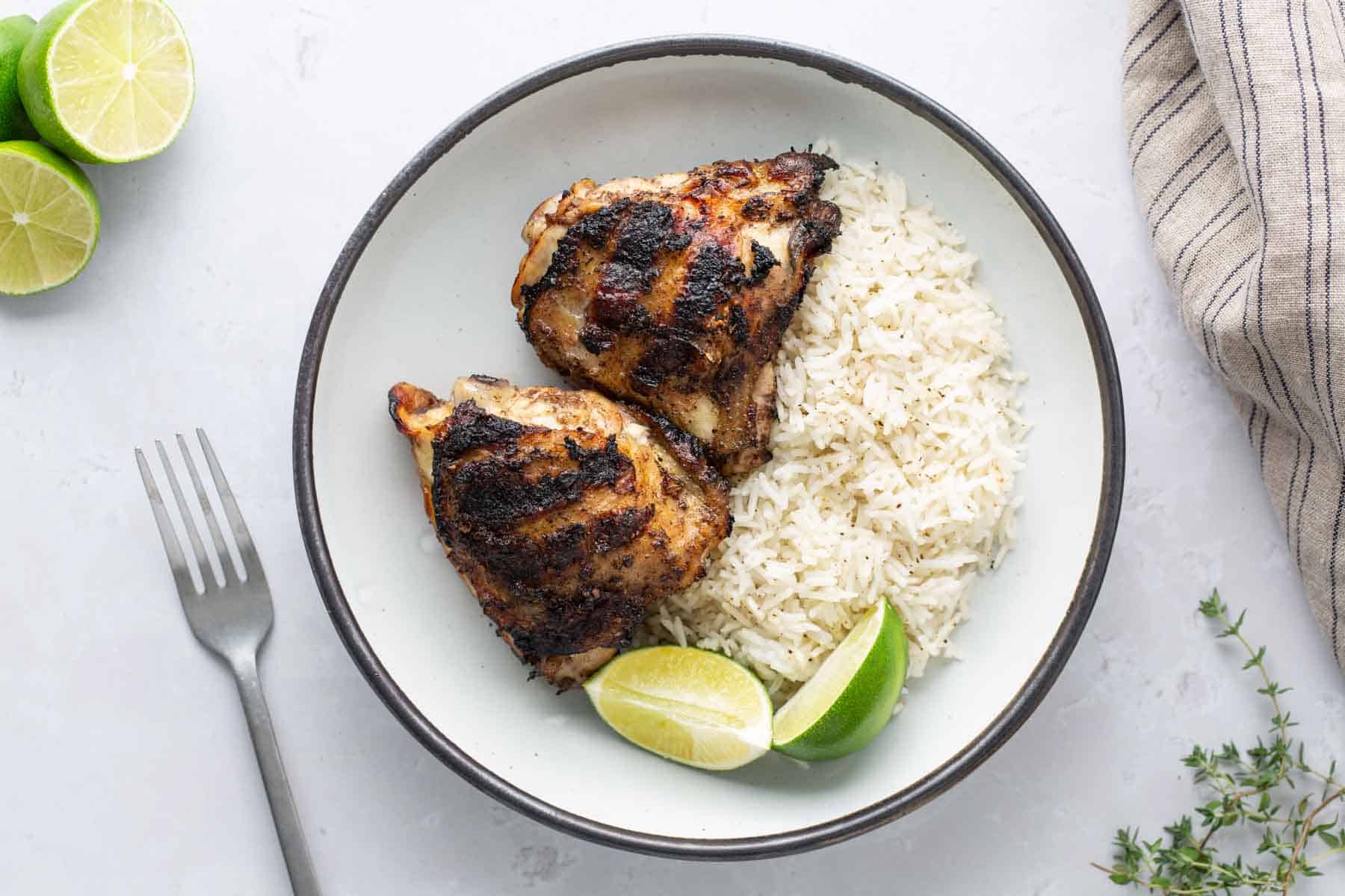 A plate with two grilled chicken thighs, white rice, and lime wedges. A fork and a striped cloth napkin are beside the plate.