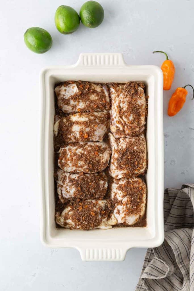 A rectangular white baking dish with seasoned chicken pieces is placed on a light surface. Three limes and two orange peppers are next to it, along with a gray cloth.