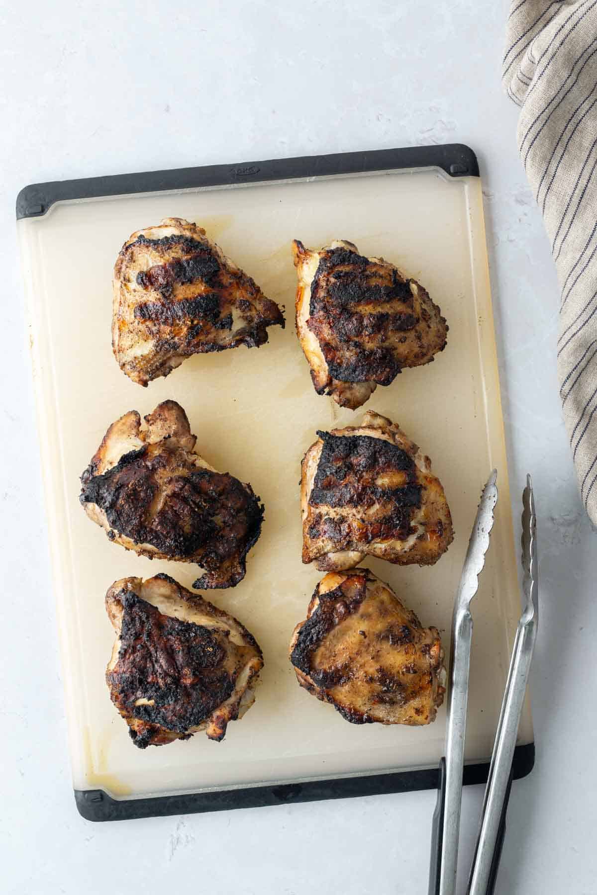 Six grilled chicken thighs on a cutting board with a pair of metal tongs and a striped cloth nearby.