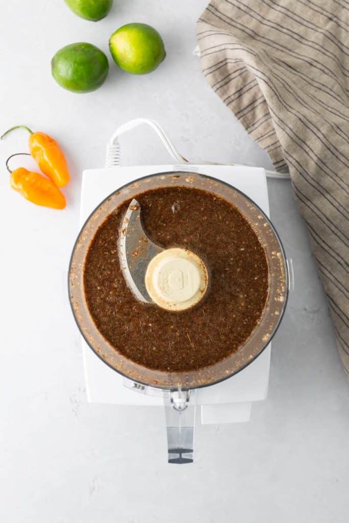 A food processor filled with blended dark brown paste sits on a countertop. Nearby are two orange habanero peppers, three limes, and a beige-striped cloth.