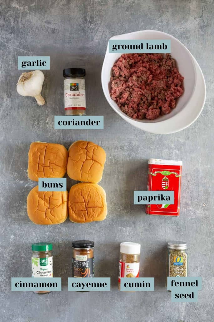 Ingredients for a recipe include ground lamb, garlic, coriander, buns, paprika, cinnamon, cayenne, cumin, and fennel seed, all laid out on a gray surface.