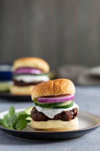 A burger with a beef patty, sliced cucumber, red onion, and a dollop of white sauce served on a plate, with another burger blurred in the background.