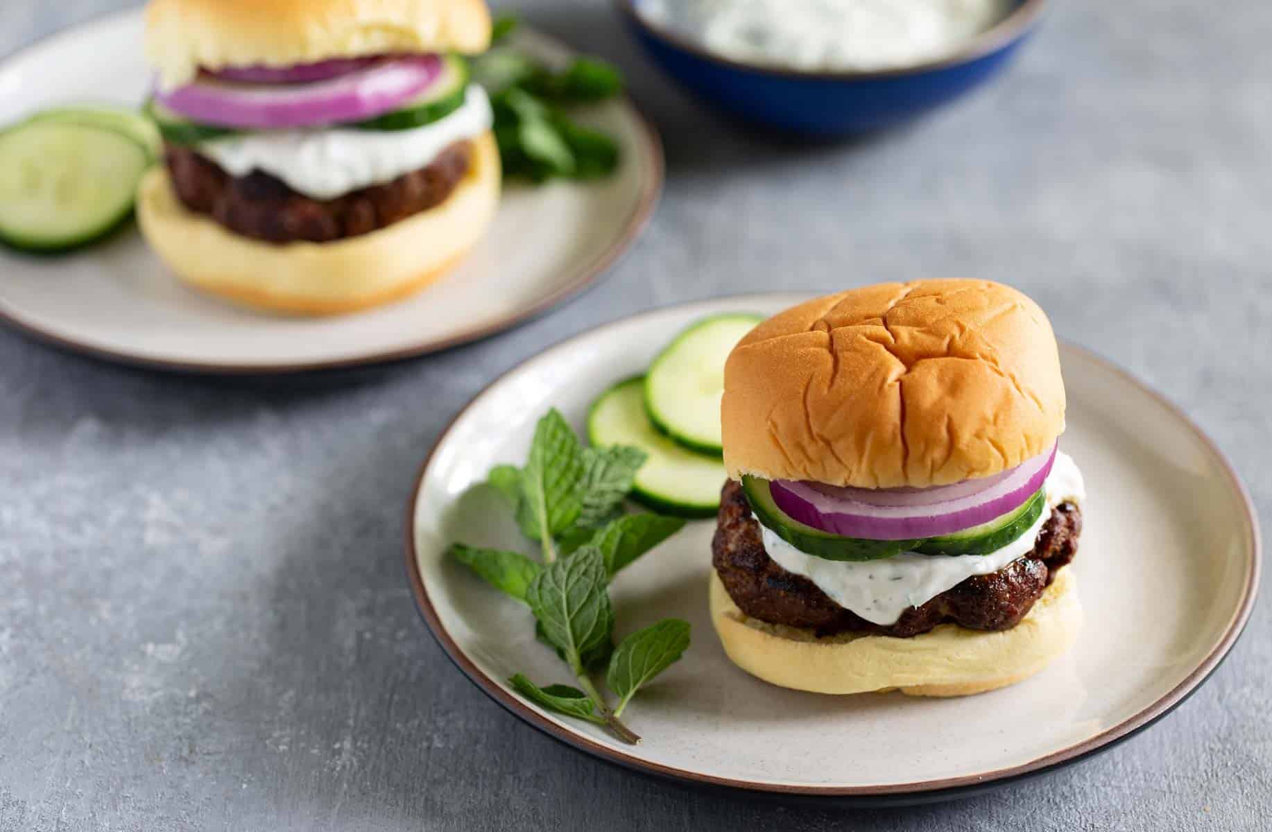 Two plates with lamb burgers topped with creamy tzatziki sauce, red onion slices, and cucumber rounds. A sprig of fresh mint and more cucumber slices accompany the burgers, with a dipping sauce in the background.