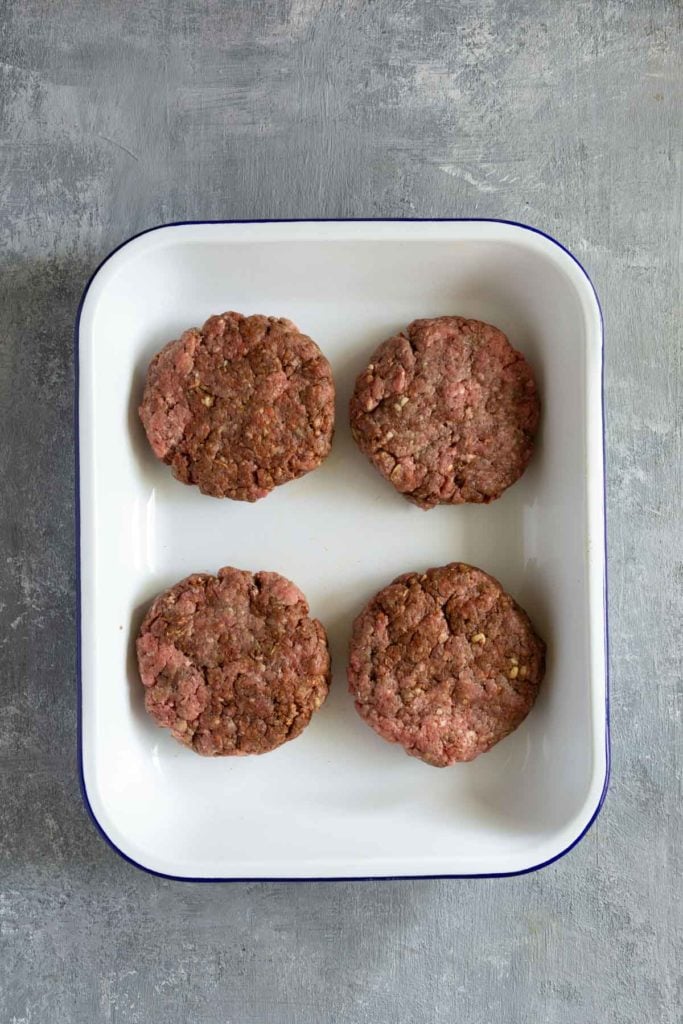 Four raw hamburger patties are arranged in a white rectangular dish with a blue rim, set on a gray textured surface.