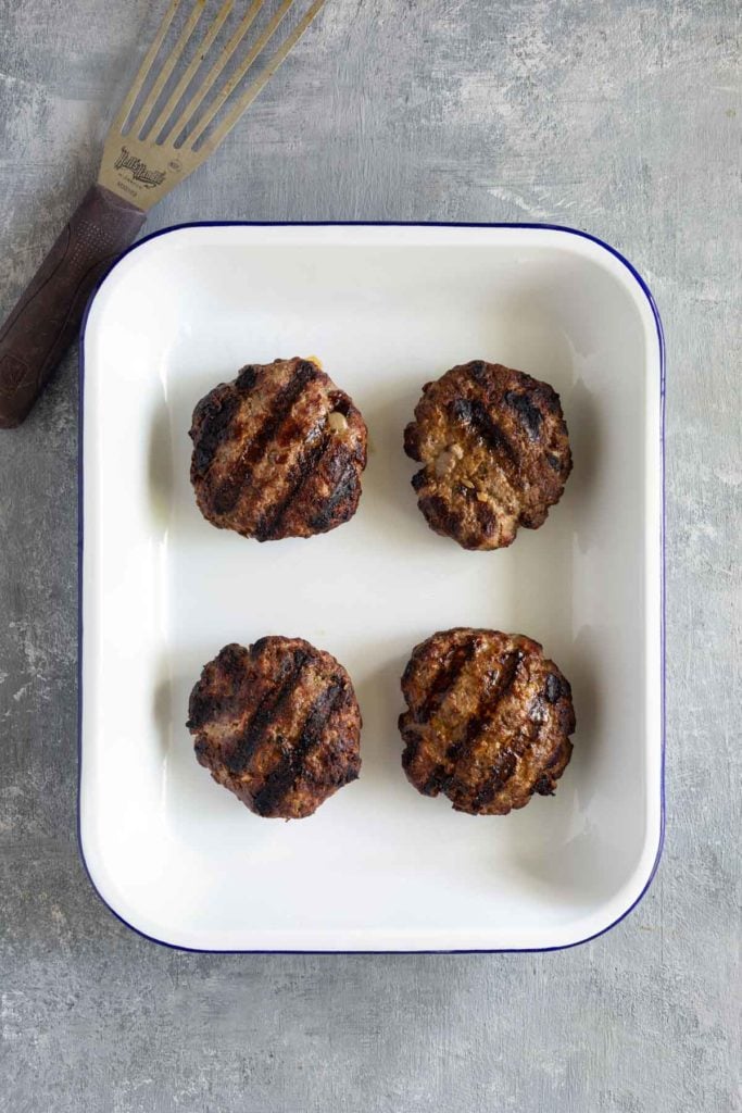 Four grilled burger patties are arranged in a white square dish with a blue rim. A grilling spatula is partially visible in the top left corner.
