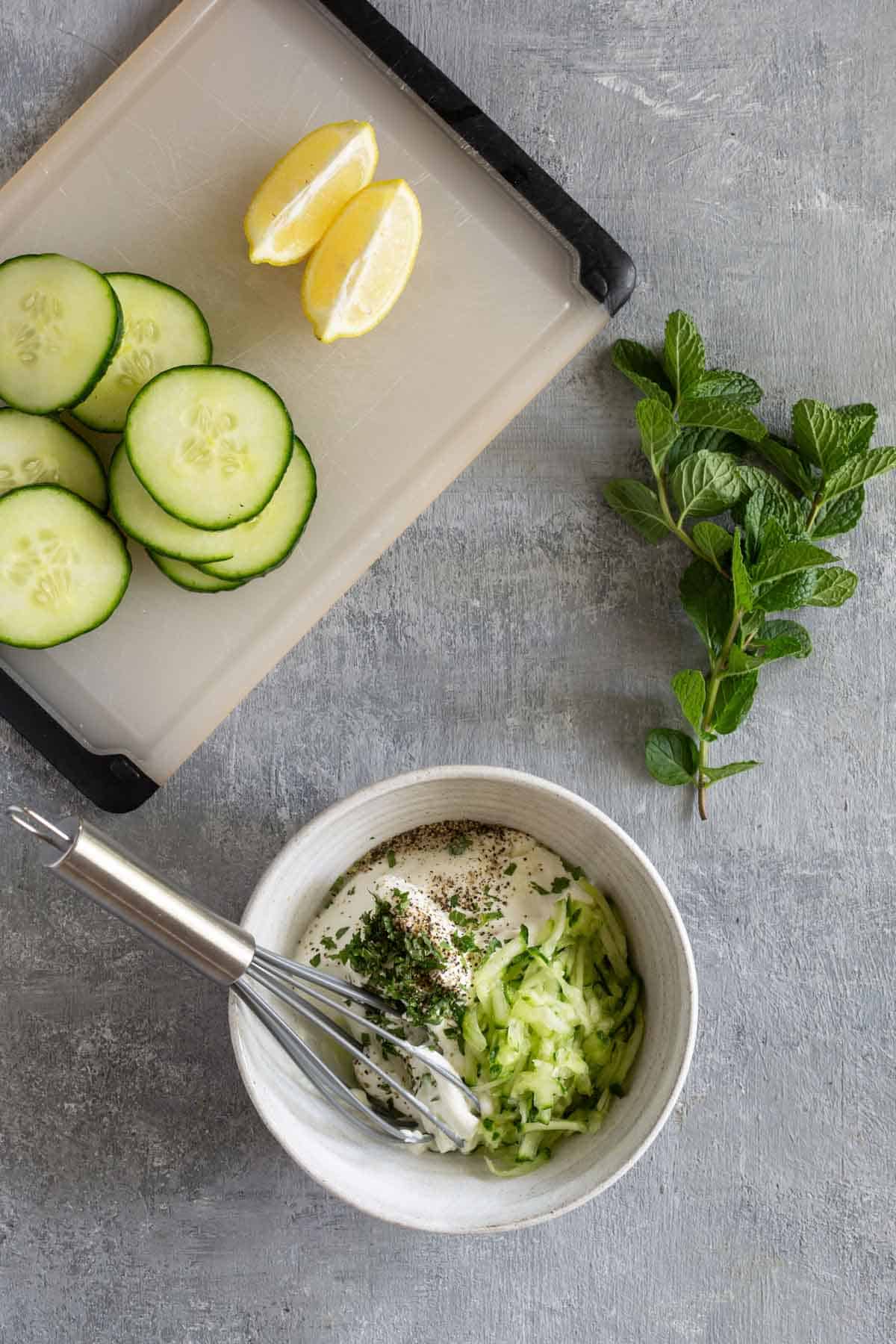 A cutting board with sliced cucumbers and lemon wedges is next to a bowl containing yogurt, chopped cucumber, and herbs, beside a whisk and fresh mint leaves on a gray surface.