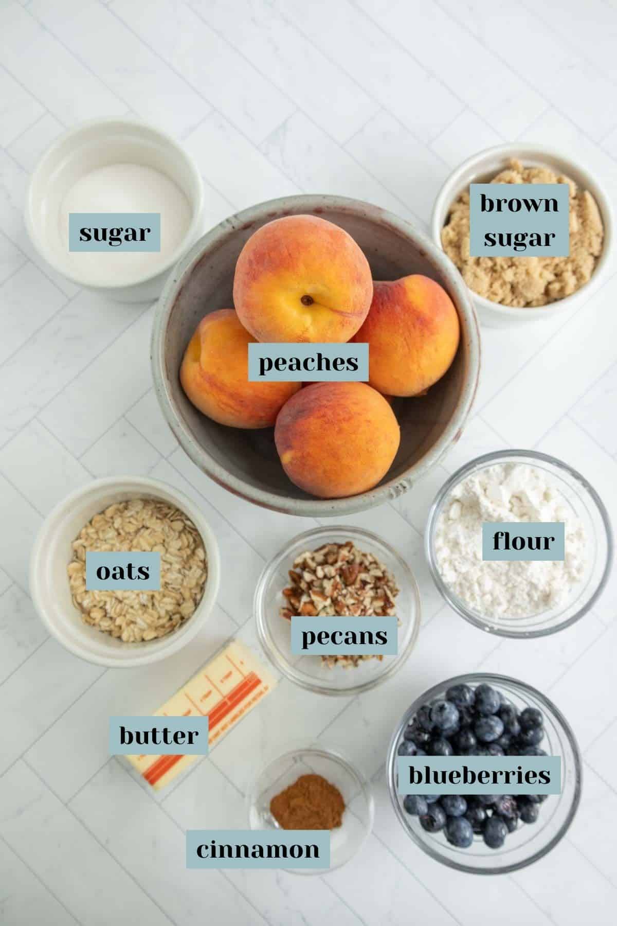 Overhead view of baking ingredients including peaches, sugar, brown sugar, oats, pecans, flour, butter, cinnamon, and blueberries in bowls on a white surface. Each ingredient is labeled.