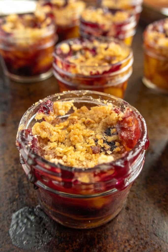 Close-up of a small glass jar filled with fruit crumble dessert, with more jars of the same dessert in the background on a dark surface.