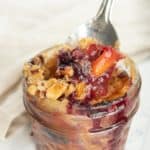 Close-up of a spoon scooping a portion of peach and berry crumble from a small glass jar. The crumble has a golden-brown topping with visible pieces of fruit and berry juice.