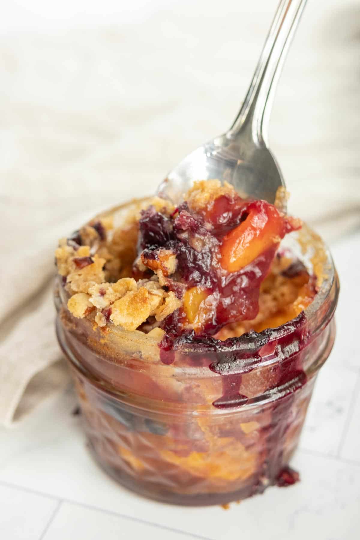 Close-up of a spoon scooping a portion of peach and berry crumble from a small glass jar. The crumble has a golden-brown topping with visible pieces of fruit and berry juice.