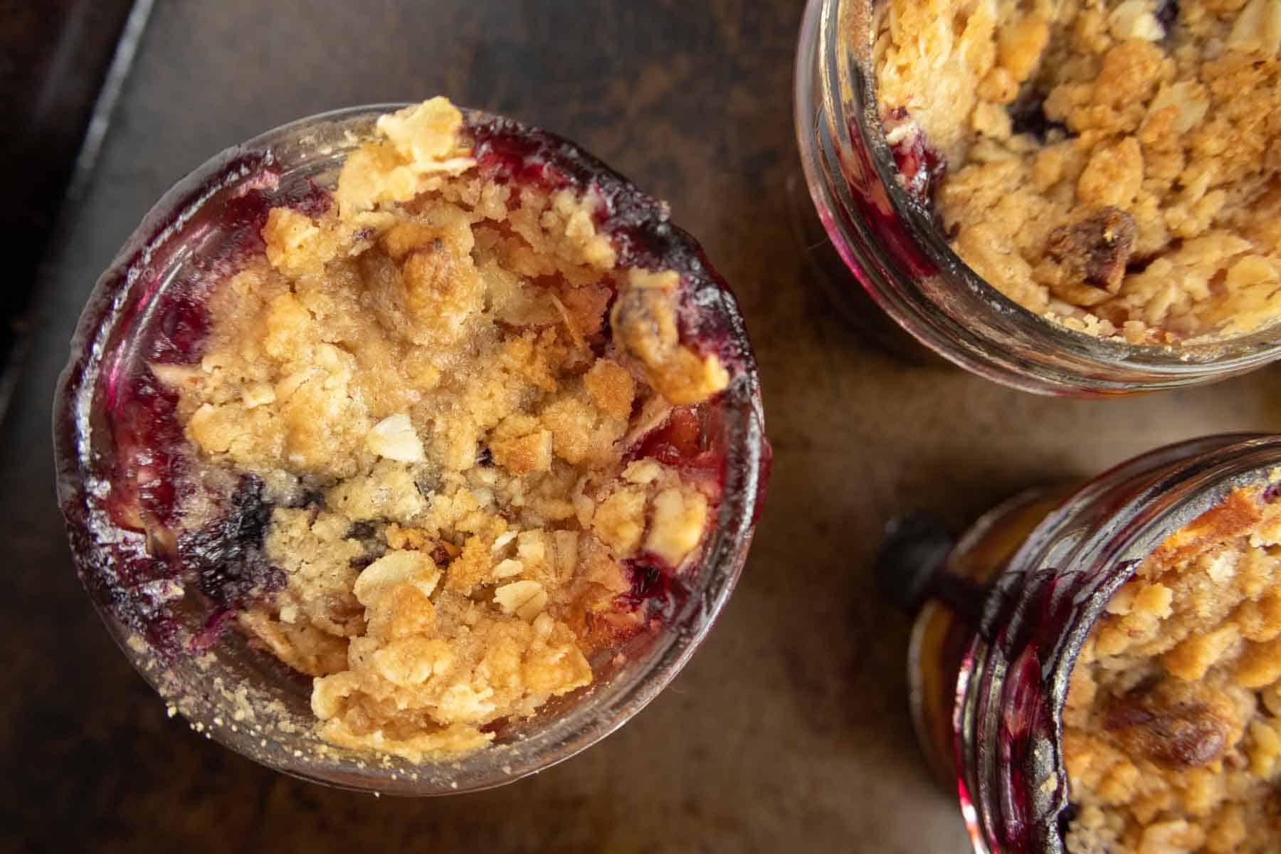 Top view of three small jars filled with berry crumble topped with a golden oat and nut crust on a dark surface.