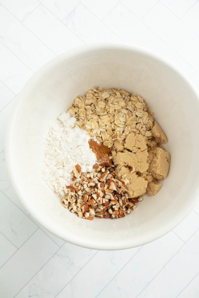 A white bowl containing oatmeal, flour, chopped nuts, brown sugar, and a small amount of cinnamon.