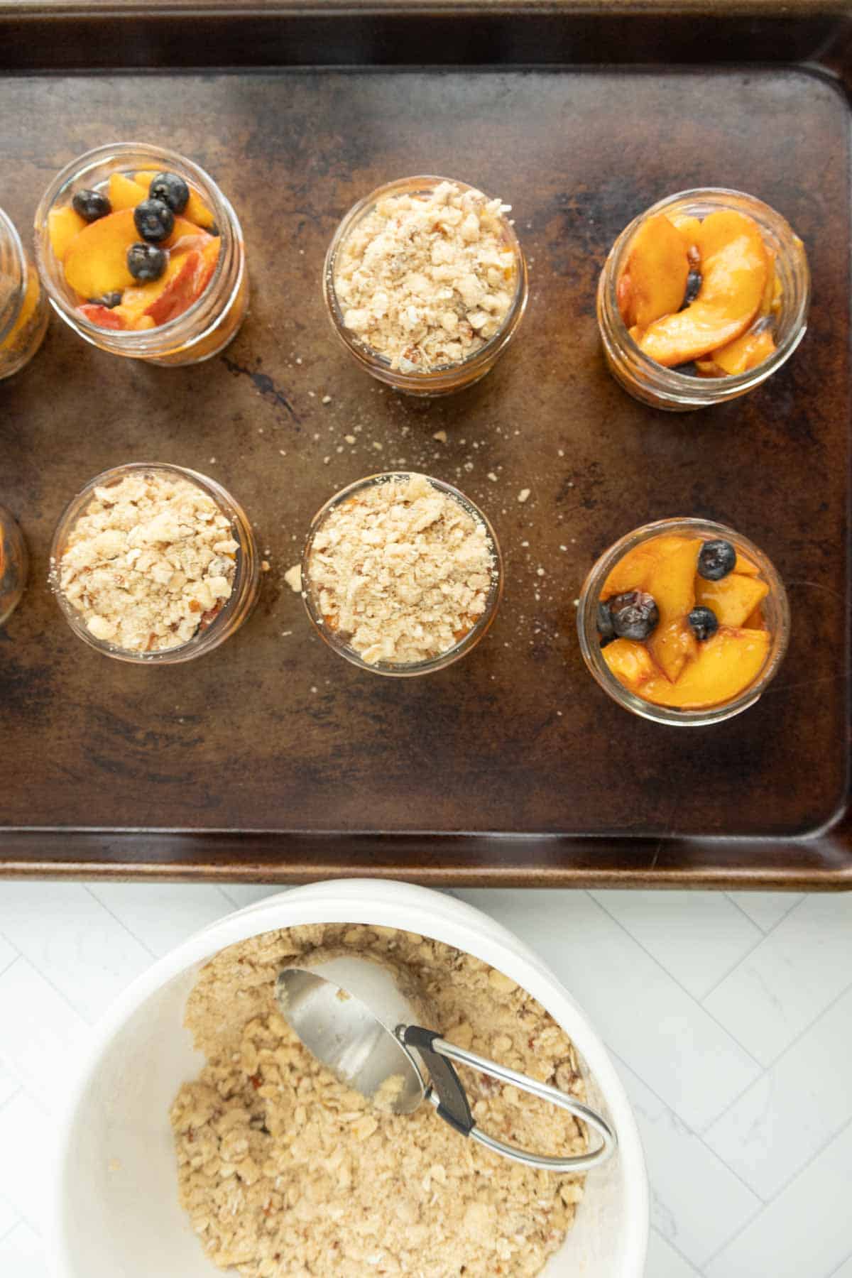 A baking tray with six jars of fruit crisps, some topped with fresh fruit and others with a crumble topping. A bowl with crumble mixture and a scoop is placed below the tray.