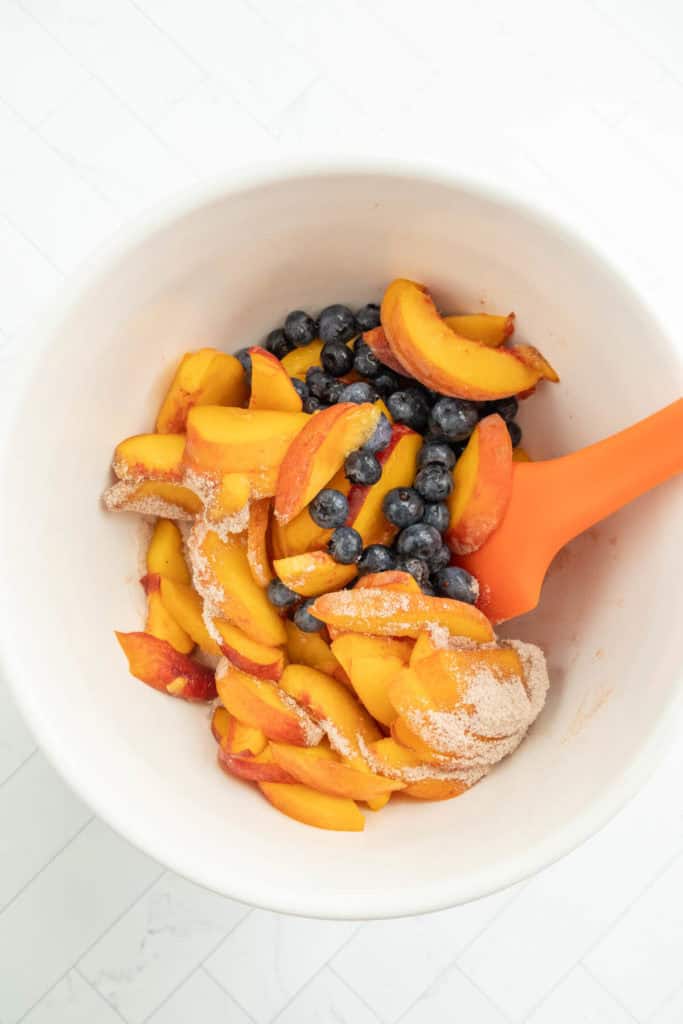 A white bowl contains sliced peaches, blueberries, and a mixture of sugar and spices being mixed with an orange spatula.