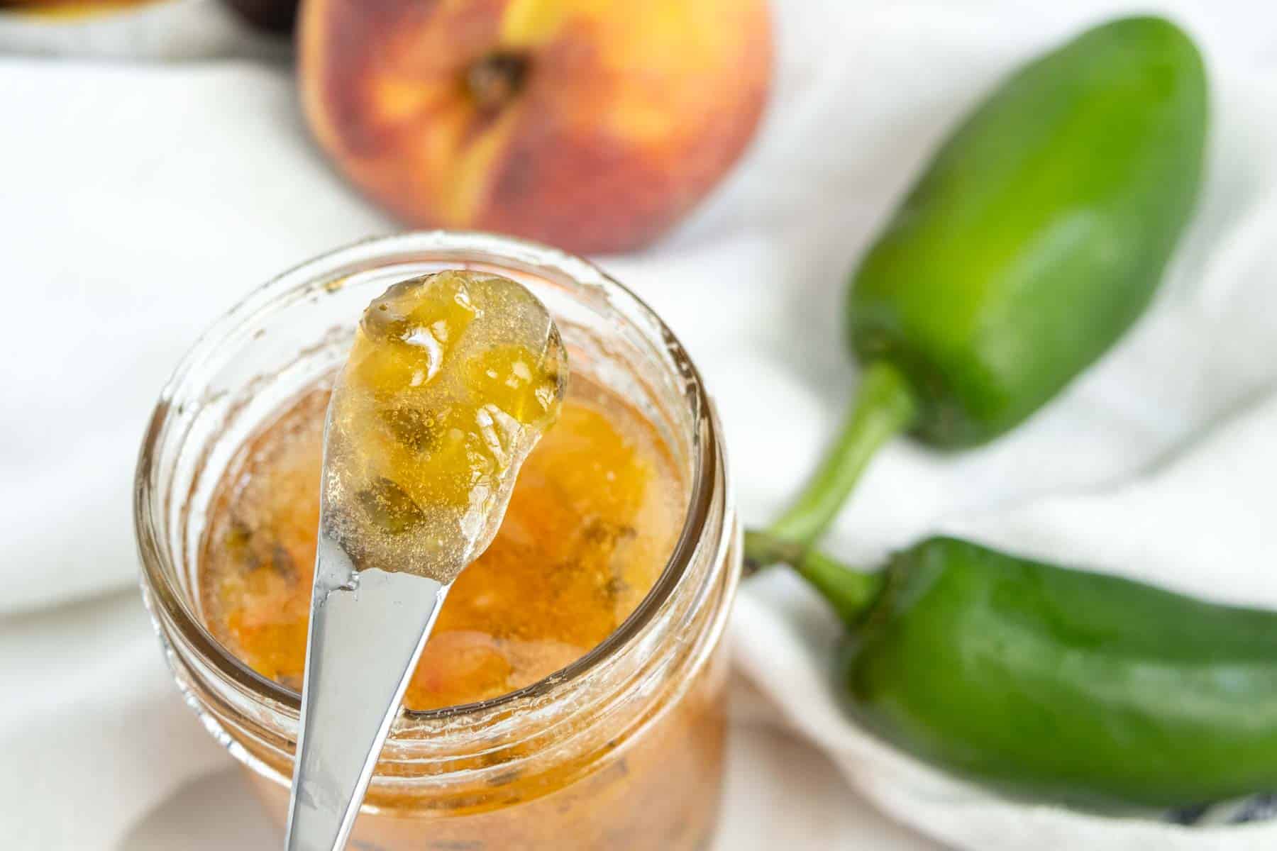 Close-up of a spoonful of yellowish jam being held above an open jar. In the background are a peach and two green chili peppers on a white cloth.