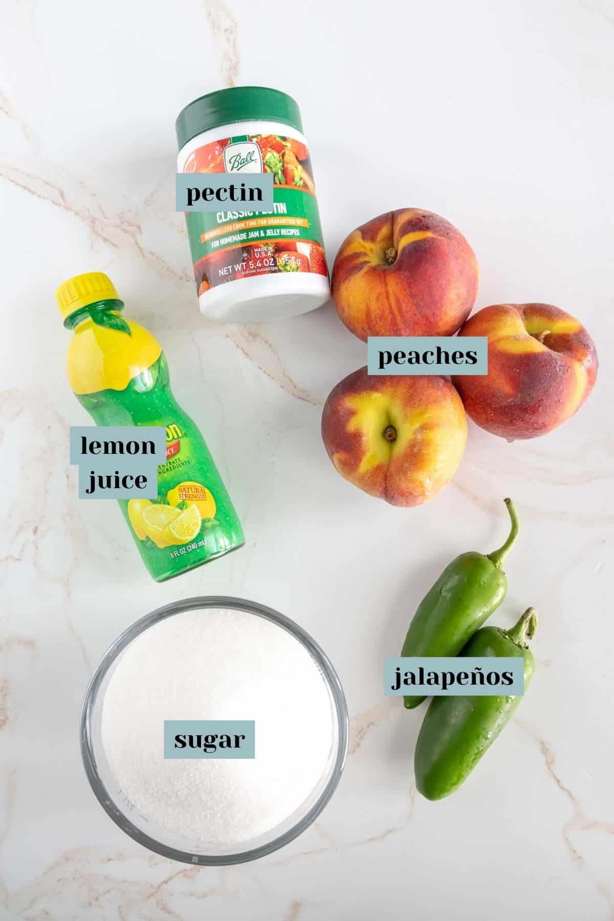 Ingredients for a recipe are arranged on a surface; pectin, peaches, lemon juice, jalapeños, and a bowl of sugar, each labeled clearly.