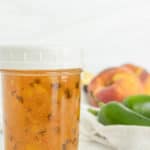 A jar of homemade peach jalapeño jam sits on a counter. In the background, fresh peaches and jalapeños are visible.