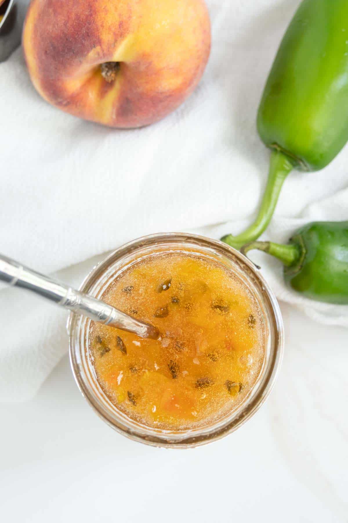 A jar of peach jalapeño jam is surrounded by fresh peaches and jalapeños on a white cloth. A spreader is inside the jar.