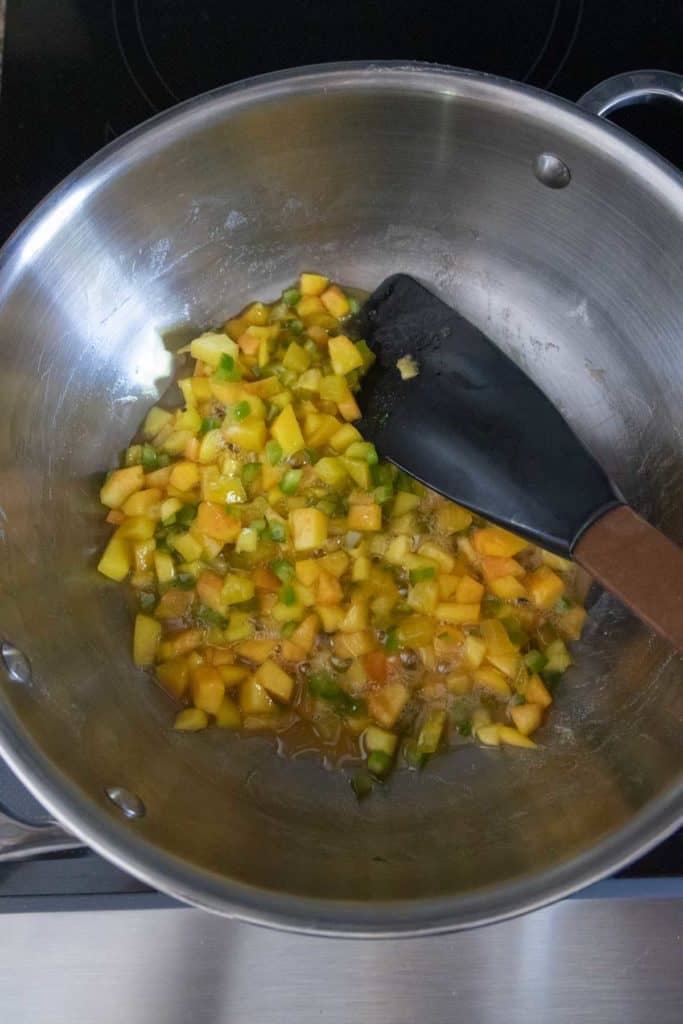 A mixture of diced fruits and vegetables being cooked in a stainless steel pan, stirred with a black spatula.