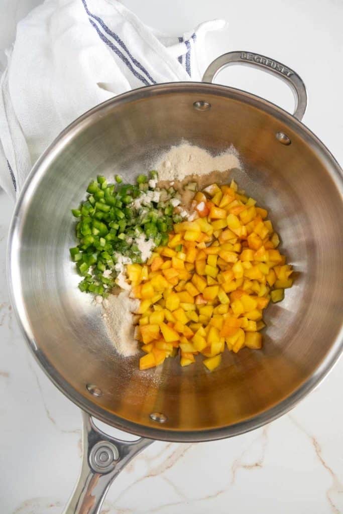 Stainless steel bowl containing chopped green bell peppers, diced peaches, minced garlic, and a mix of spices. A white kitchen towel is placed beside the bowl.