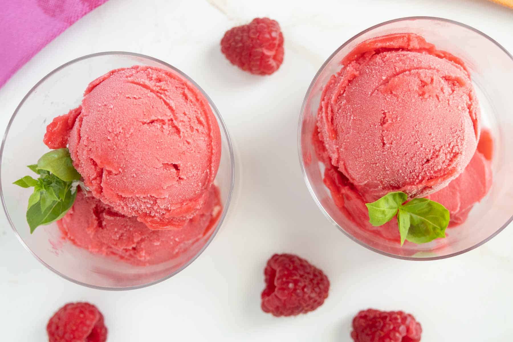 Top-down view of two bowls of raspberry sorbet garnished with basil leaves, surrounded by fresh raspberries on a white surface.