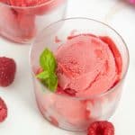 Two clear glasses filled with scoops of bright pink raspberry sorbet, garnished with fresh mint leaves, are placed on a white surface with raspberries scattered around.