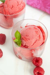 Two clear glasses filled with scoops of bright pink raspberry sorbet, garnished with fresh mint leaves, are placed on a white surface with raspberries scattered around.