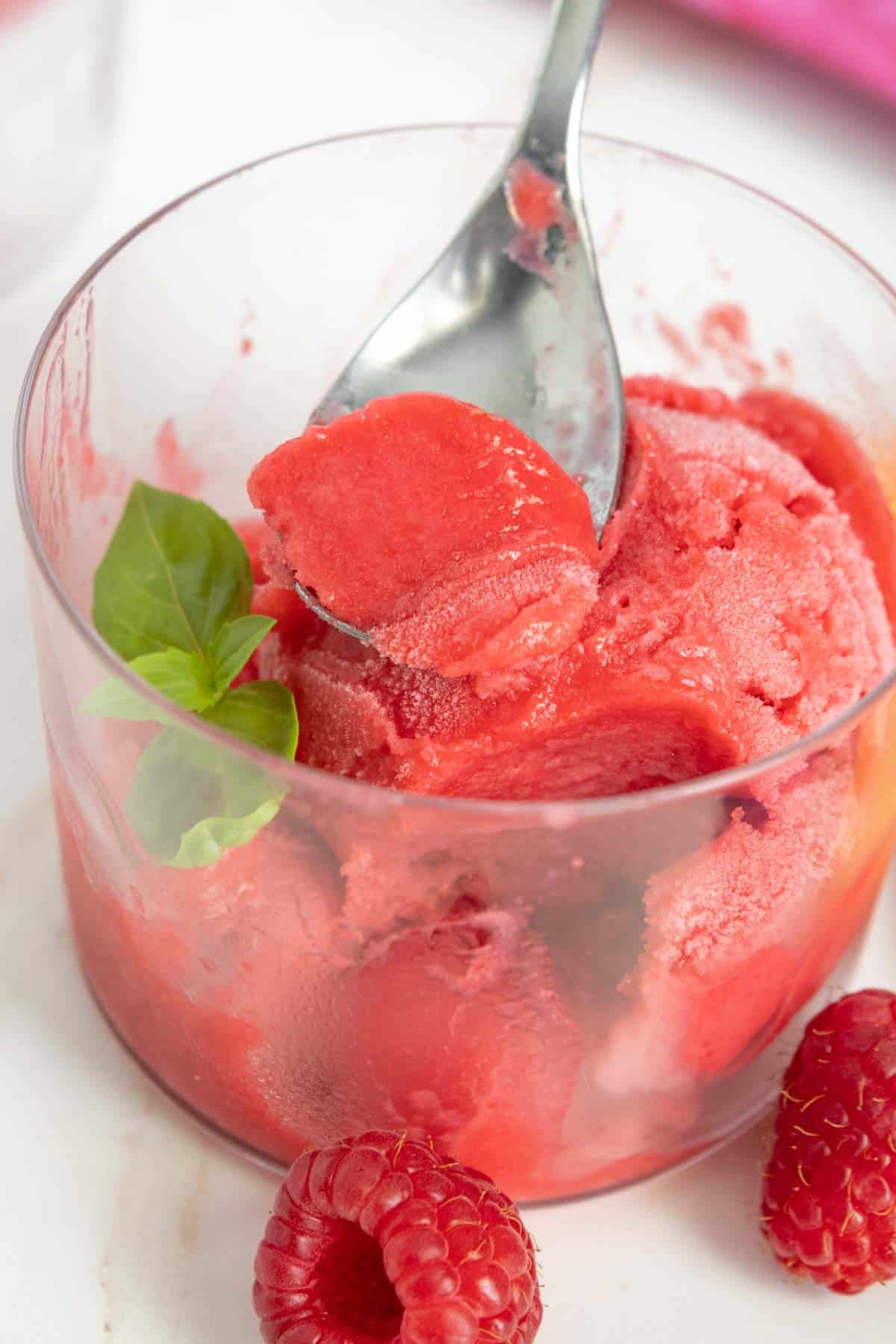 A spoon scoops vibrant red raspberry sorbet from a glass bowl, garnished with fresh basil leaves and surrounded by whole raspberries.