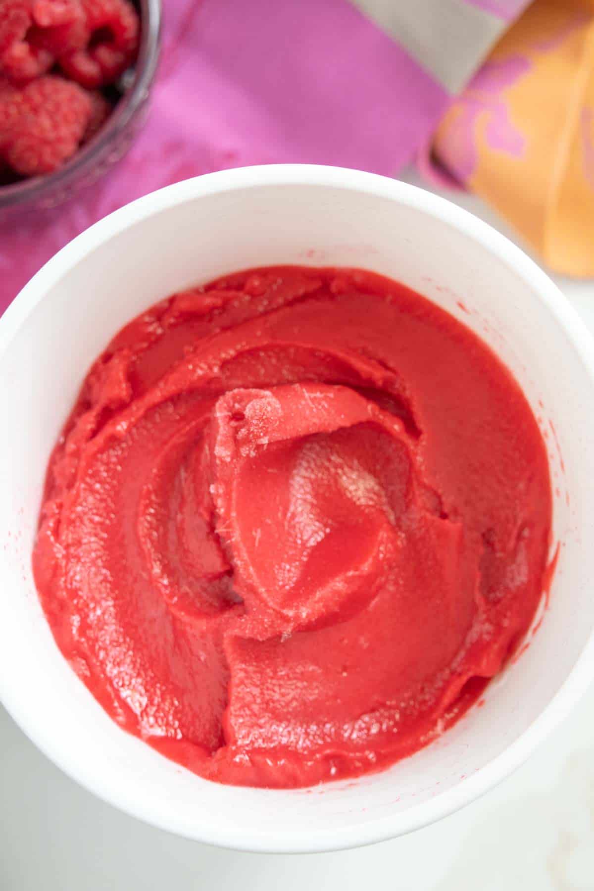 A white bowl contains smooth, bright red raspberry sorbet, with a purple cloth and a small bowl of raspberries in the background.