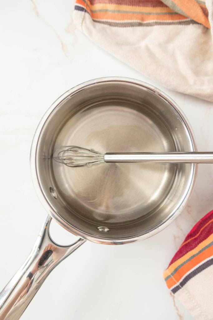 A stainless steel saucepan filled with water and a whisk, placed on a white surface with a striped towel beside it.