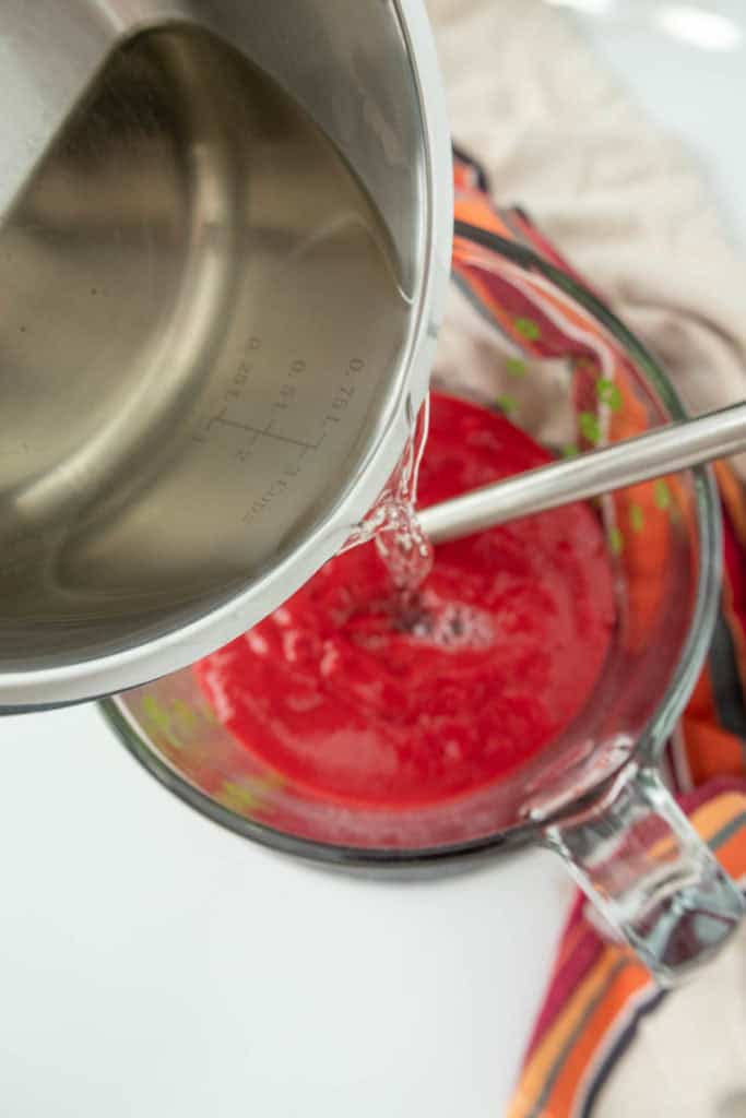 A pot of liquid is being poured into a measuring cup filled with a red mixture, with a whisk resting inside the cup.