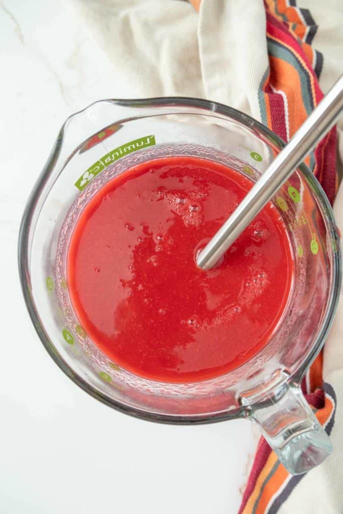 A glass measuring cup filled with red liquid, and a metal immersion blender inside it, placed on a white surface with a striped cloth in the background.