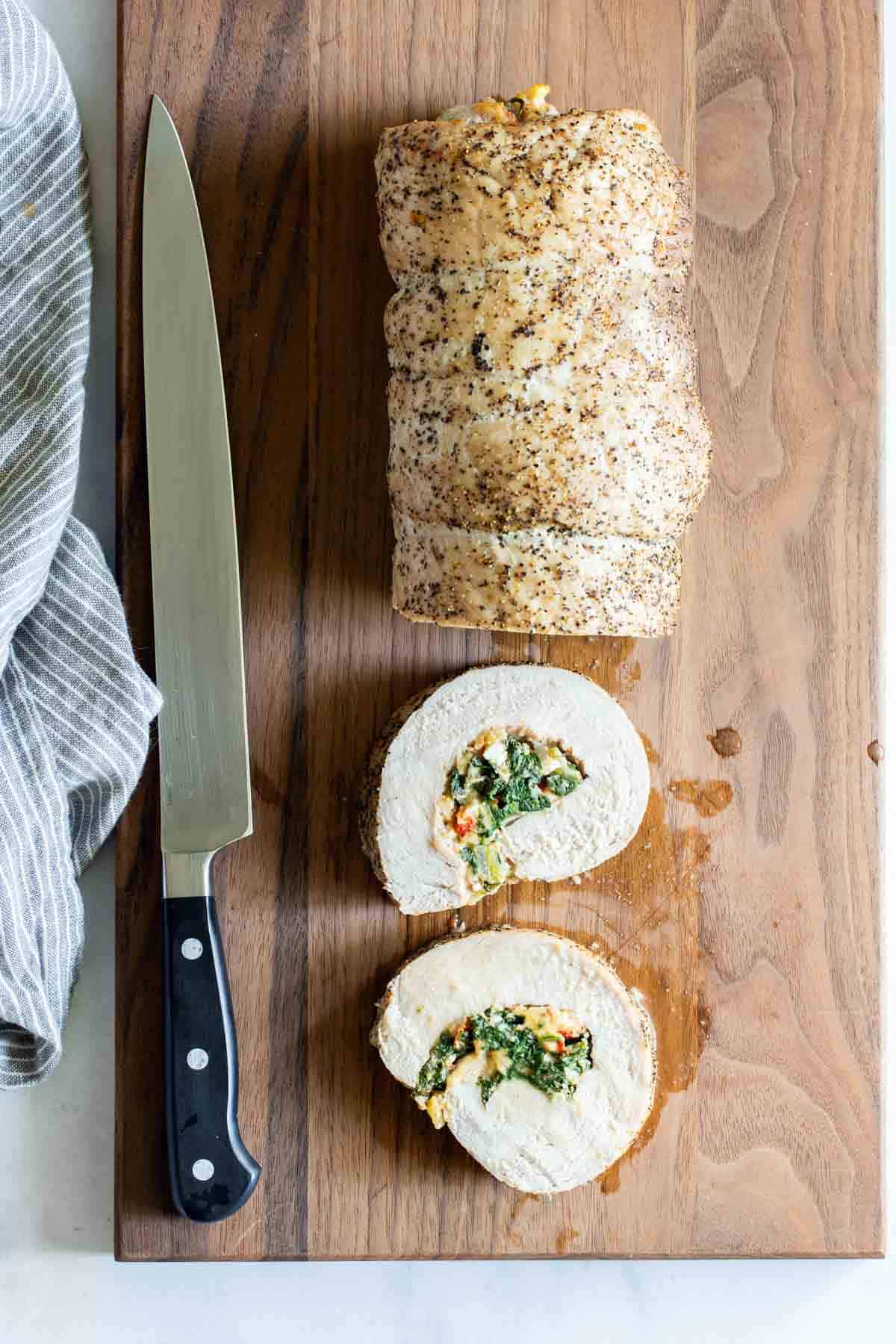 A stuffed pork roulade with two slices cut to reveal spinach and cheese filling, placed on a wooden cutting board next to a chef’s knife.