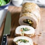 Sliced stuffed chicken roulade on a wooden cutting board with a knife on the left and a bowl of sauce in the background.