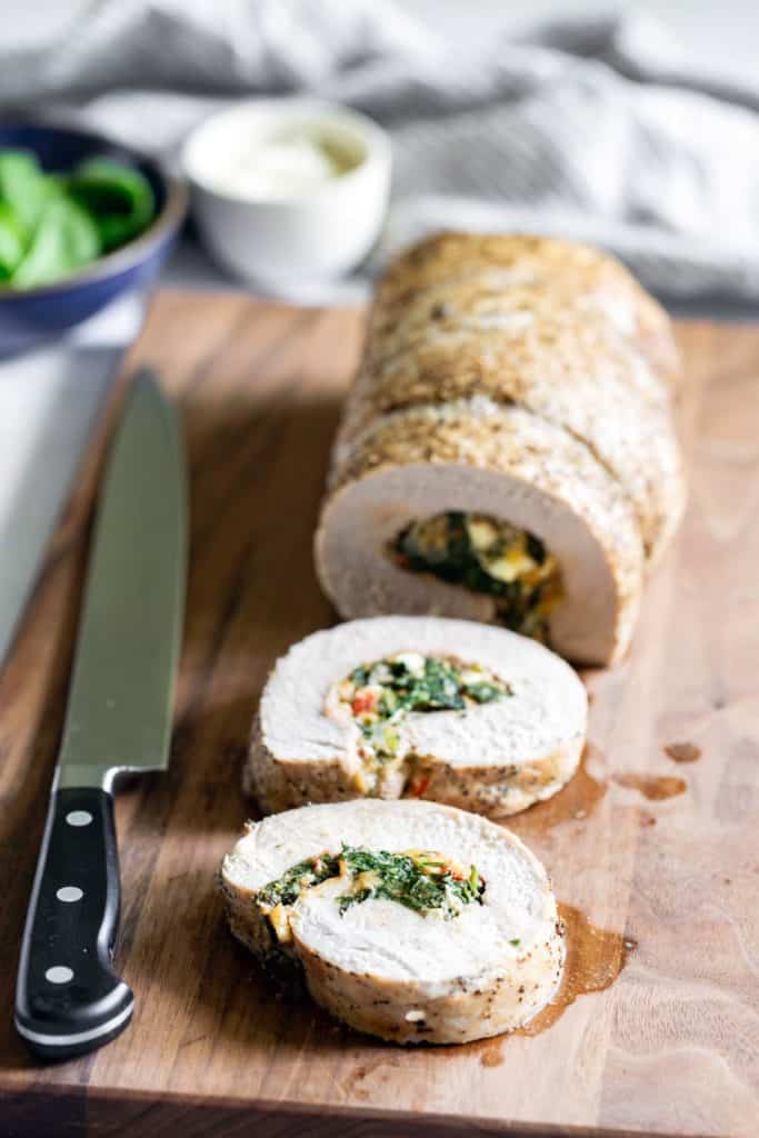 Sliced stuffed chicken roulade on a wooden cutting board with a knife on the left and a bowl of sauce in the background.