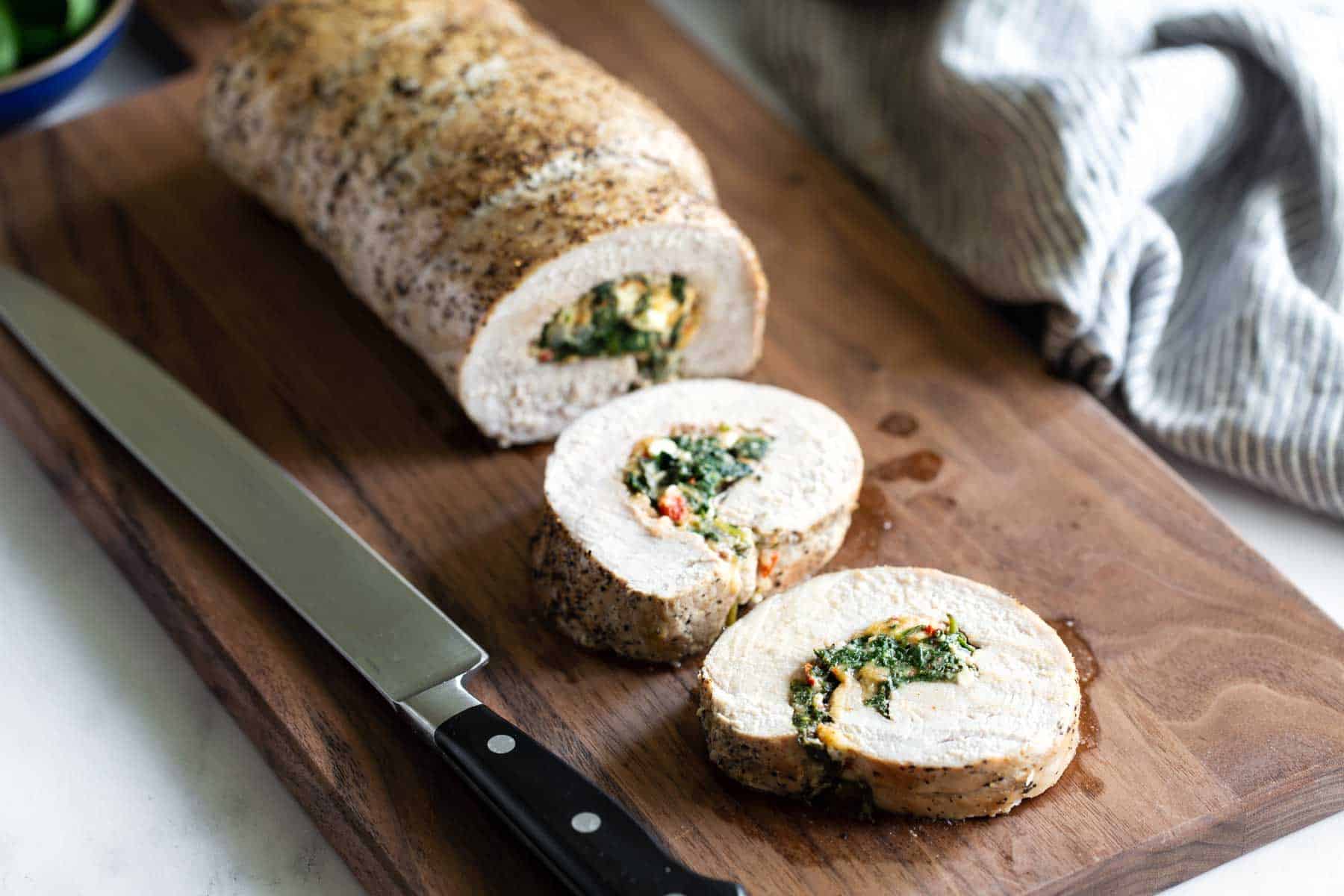 A sliced stuffed pork roll is displayed on a wooden cutting board next to a large chef's knife. The stuffing includes spinach and other ingredients. A striped cloth is in the background.