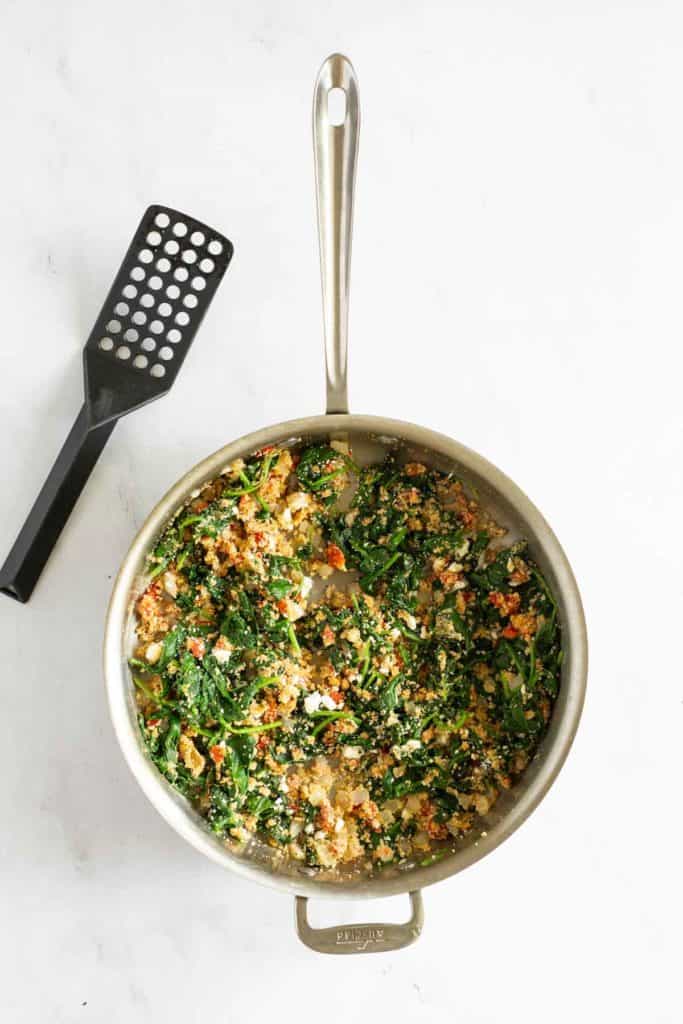 A stir-fry pan filled with cooked leafy greens and grains, accompanied by a black spatula placed next to the pan on a white surface.