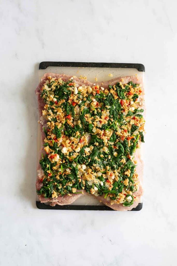 A piece of raw meat spread with a mixture of spinach, feta cheese, and chopped red peppers on a cutting board.