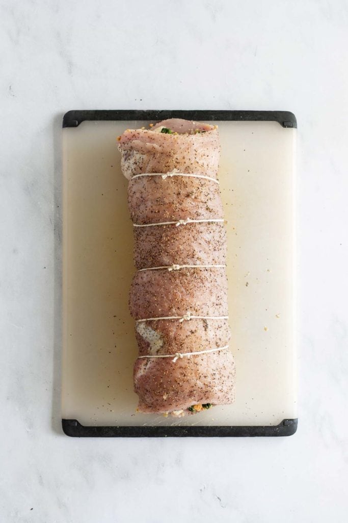 A seasoned and tied pork tenderloin is placed on a white cutting board against a white background.