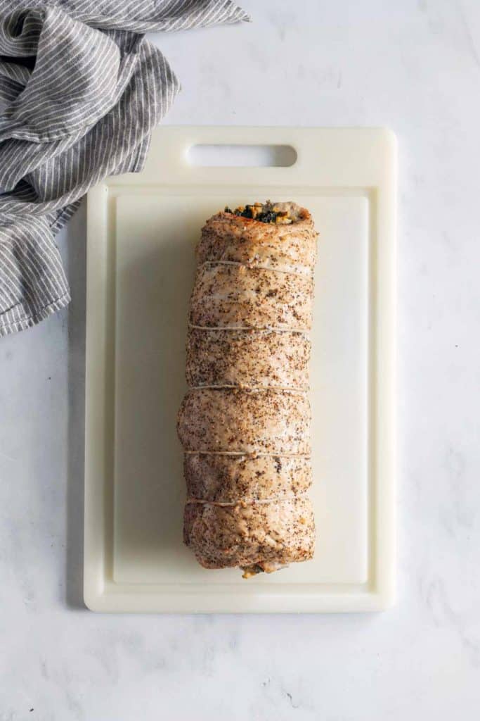 A seasoned, rolled roast tied with twine, placed on a white plastic cutting board. A gray and white striped cloth is partially visible beside the cutting board.