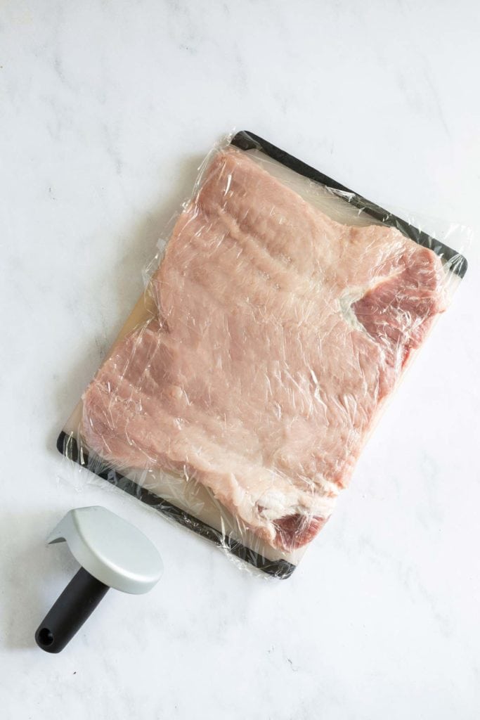 A slab of raw meat covered with plastic wrap is placed on a black tray. A meat tenderizer lies on the white surface next to the tray.