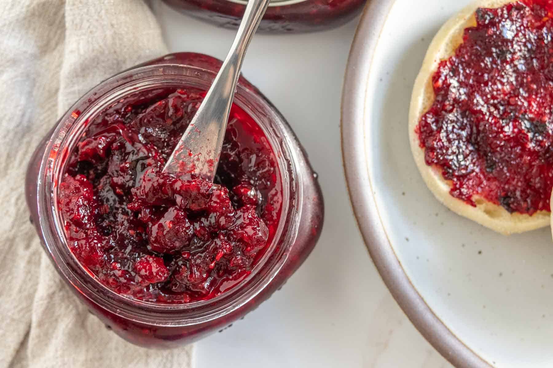 Open jar of berry jam with a spoon sticking out next to a plate holding a piece of bread spread with jam.