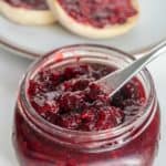 A jar of blackberry jam with a butter knife inside it, and two slices of bread spread with blackberry jam in the background.