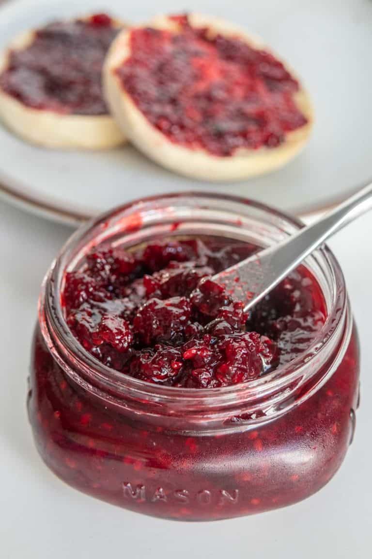 A jar of blackberry jam with a butter knife inside it, and two slices of bread spread with blackberry jam in the background.